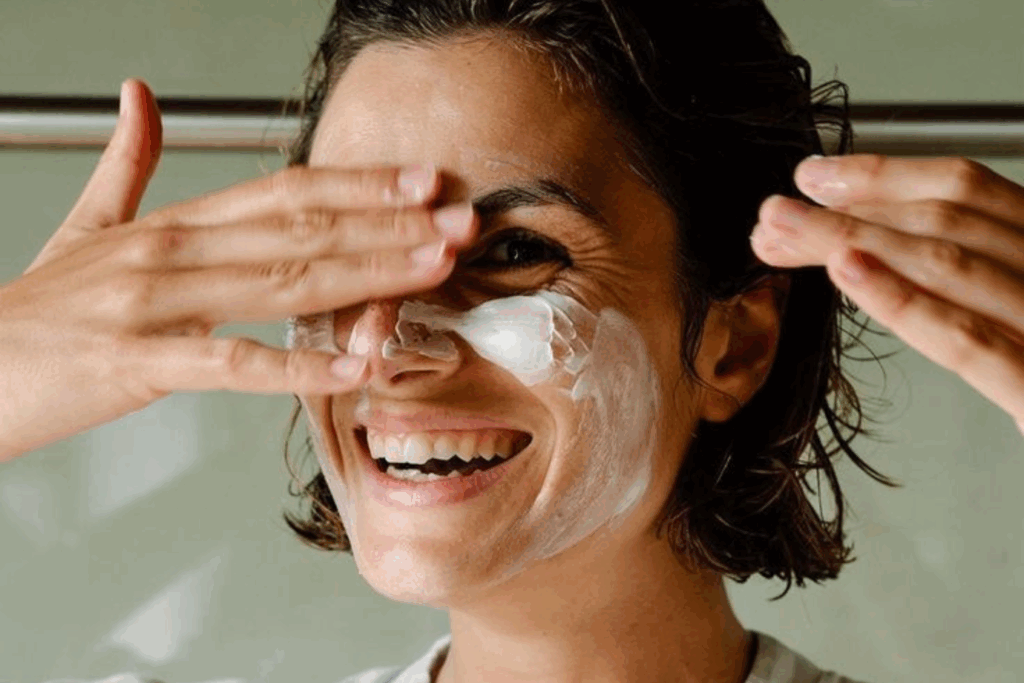 Mujer joven aplicándose crema facial con una sonrisa, en el contexto de una rutina de cuidado para prevenir el envejecimiento de la piel.